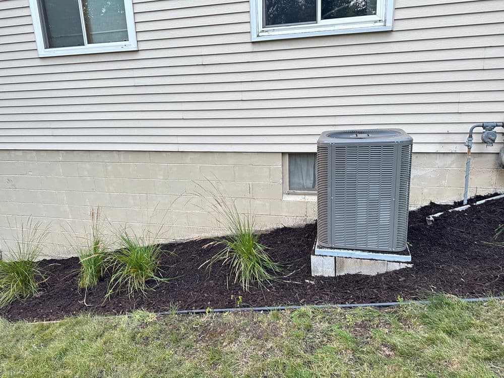 Air conditioning unit beside a house, featuring mulch and ornamental grasses in landscape design.