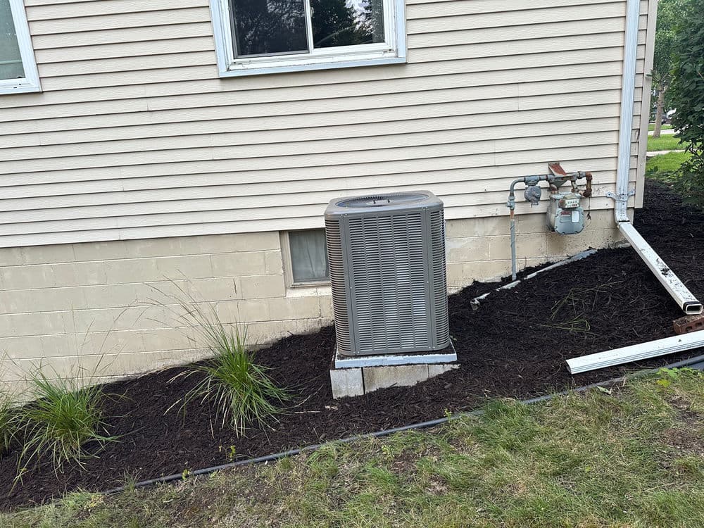 Air conditioning unit beside a house with freshly mulched landscaping and gas meter.