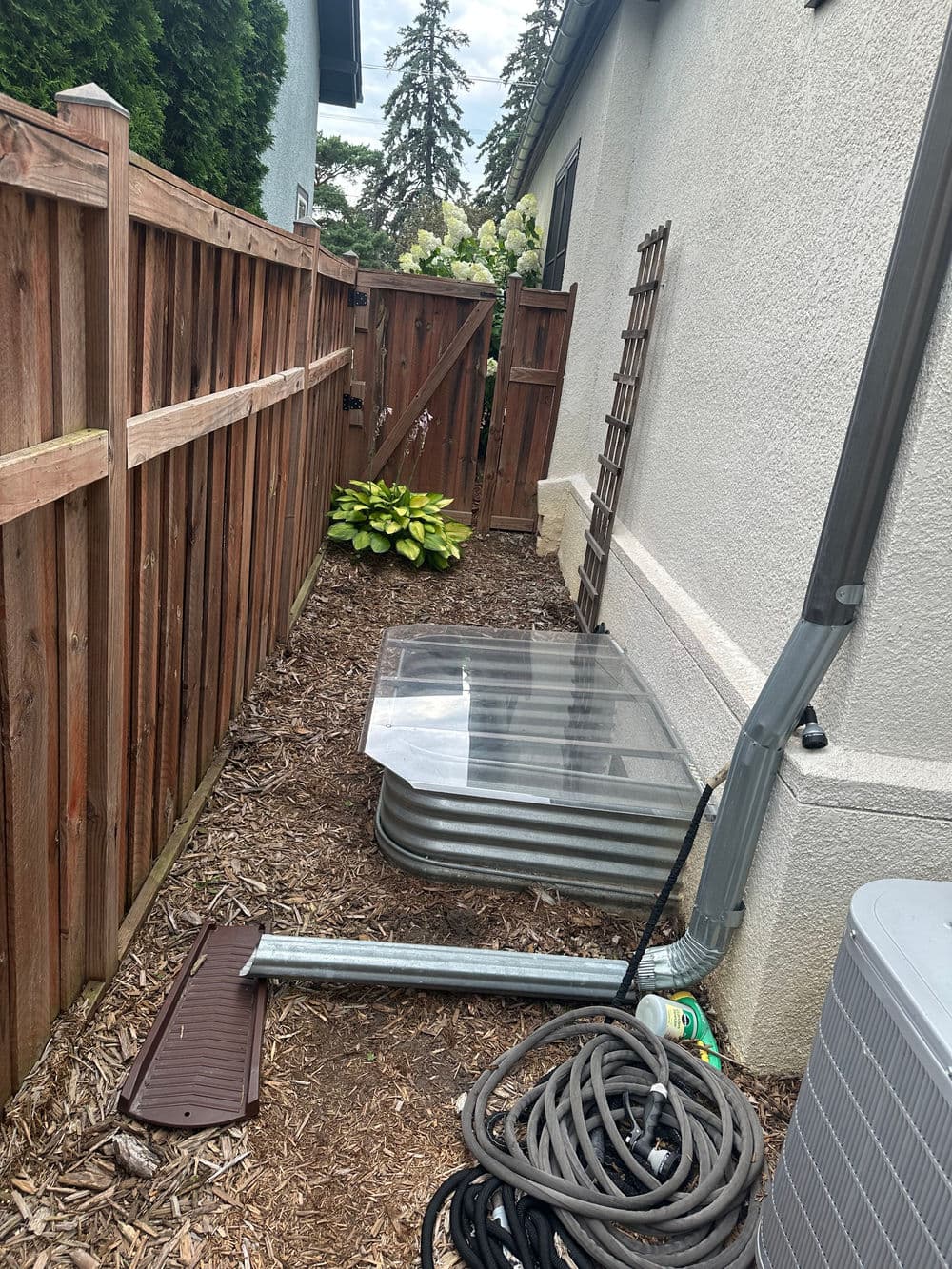 Narrow side yard with wooden fence, drainage pipes, and green plants near a house.