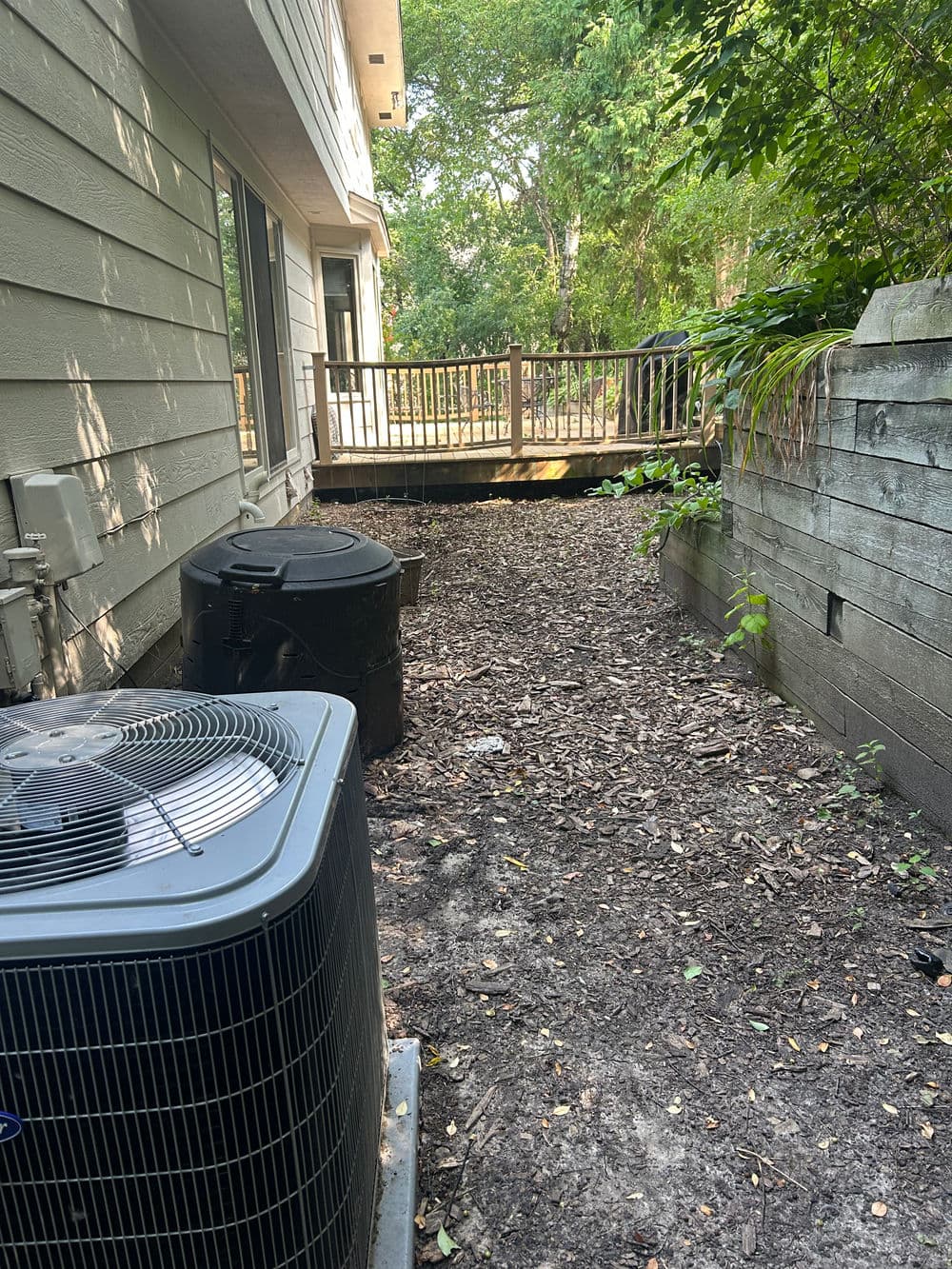 Air conditioning unit by wooded area and wooden deck, featuring a gravel and mulch pathway.