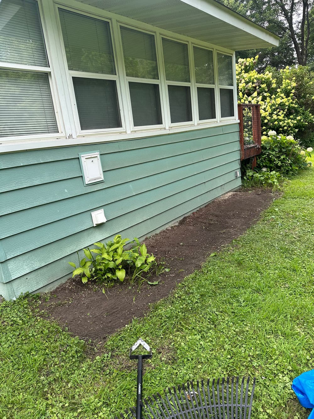 Garden bed preparation beside a house, featuring freshly turned soil and green plants.