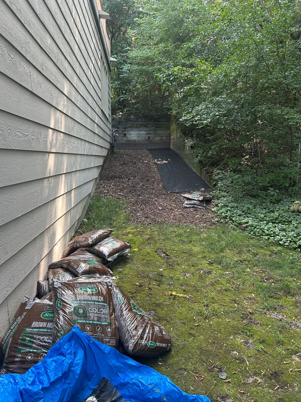 Bags of brown mulch and landscaping fabric beside a house, surrounded by greenery.
