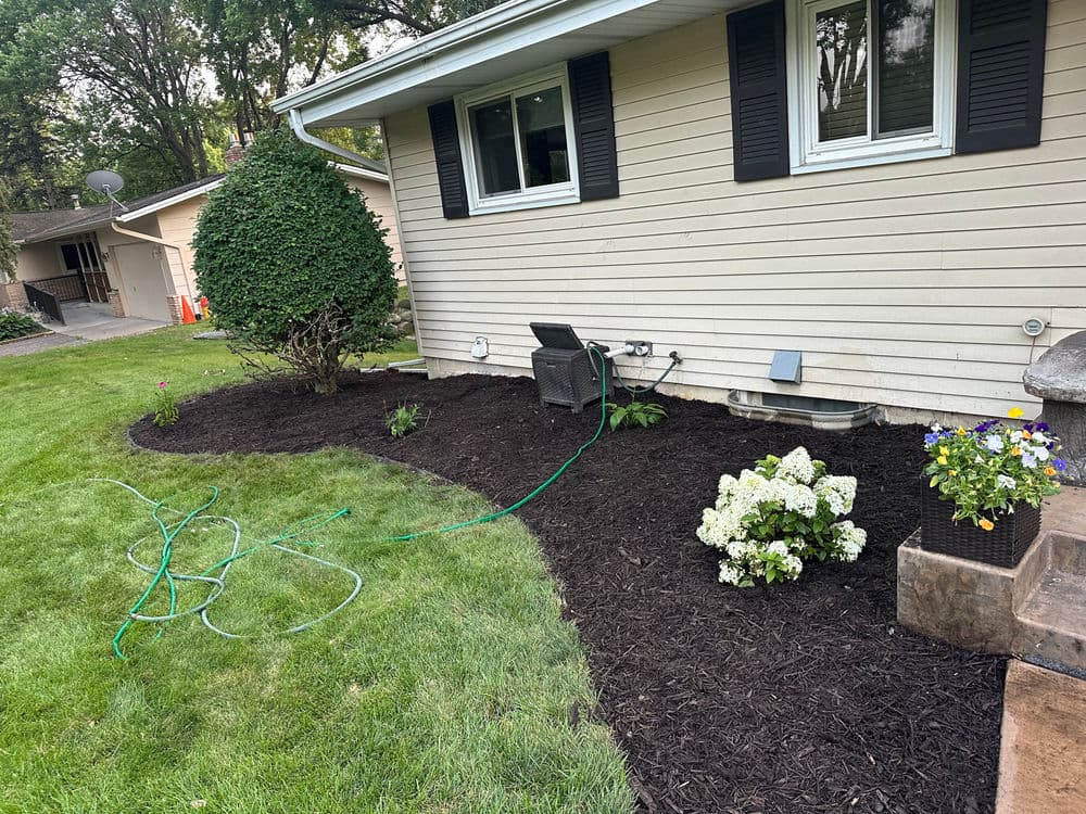 Landscaped yard with mulch, flowers, and patio furniture next to a home. Green grass and garden hose visible.