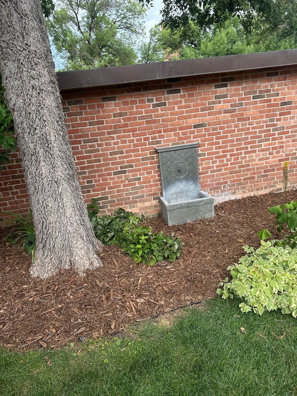 Stone water fountain near a brick wall, surrounded by greenery and mulch in a garden setting.