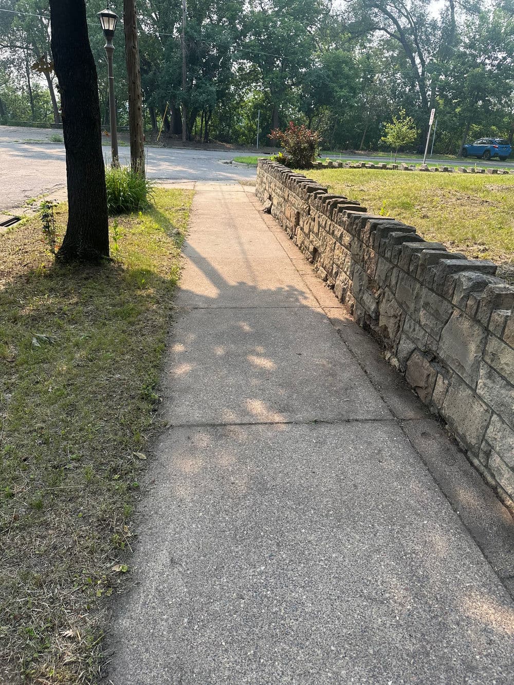 Sidewalk view beside a stone wall, surrounded by greenery and trees, on a sunny day.