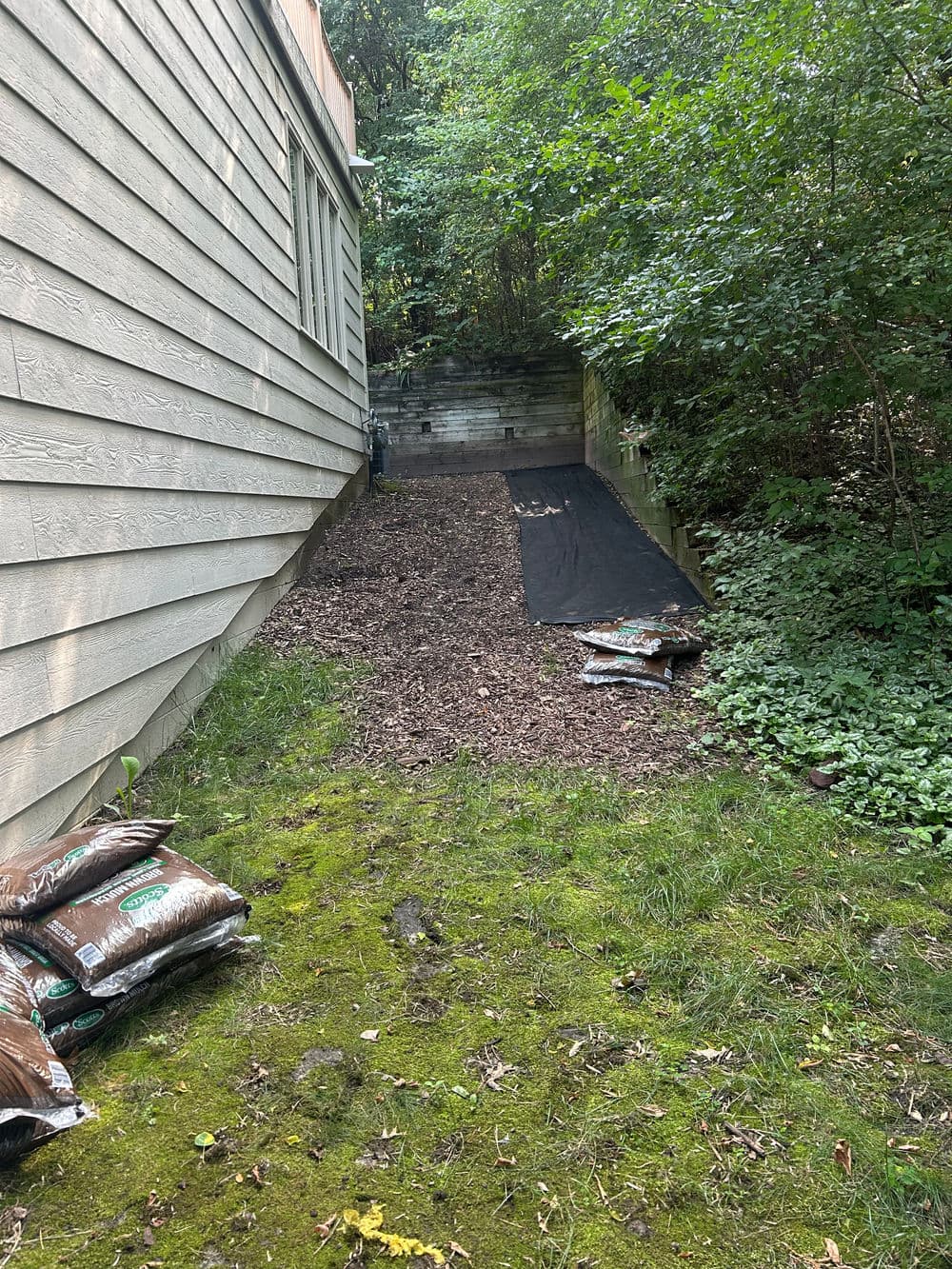 Mulched garden area beside a house, featuring landscape fabric and bags of soil.