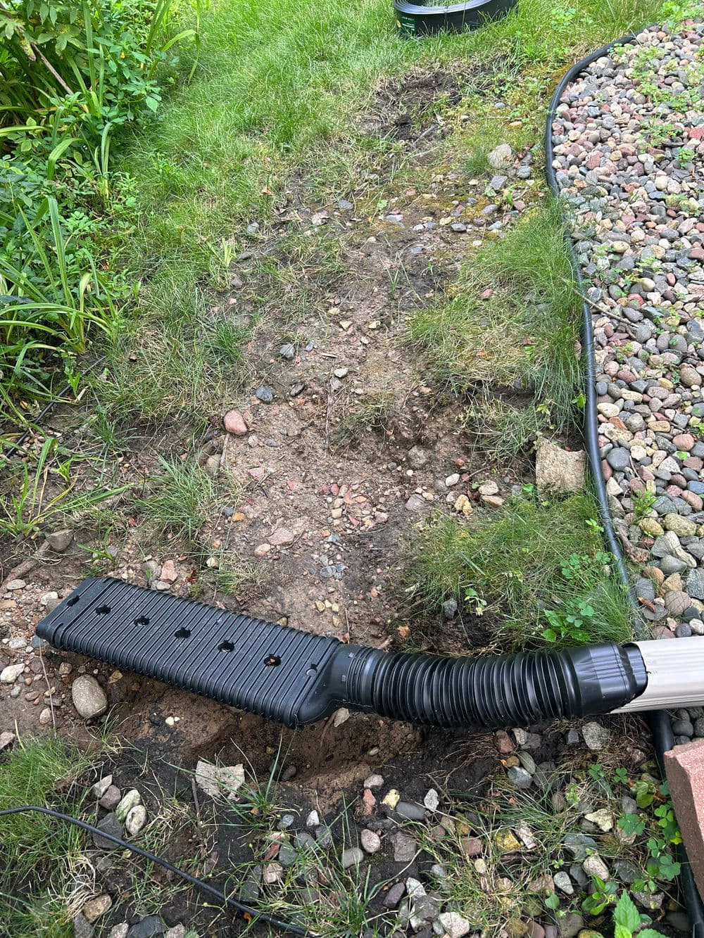 Landscape drain cover lying on gravel, surrounded by grass and dirt, ready for installation.