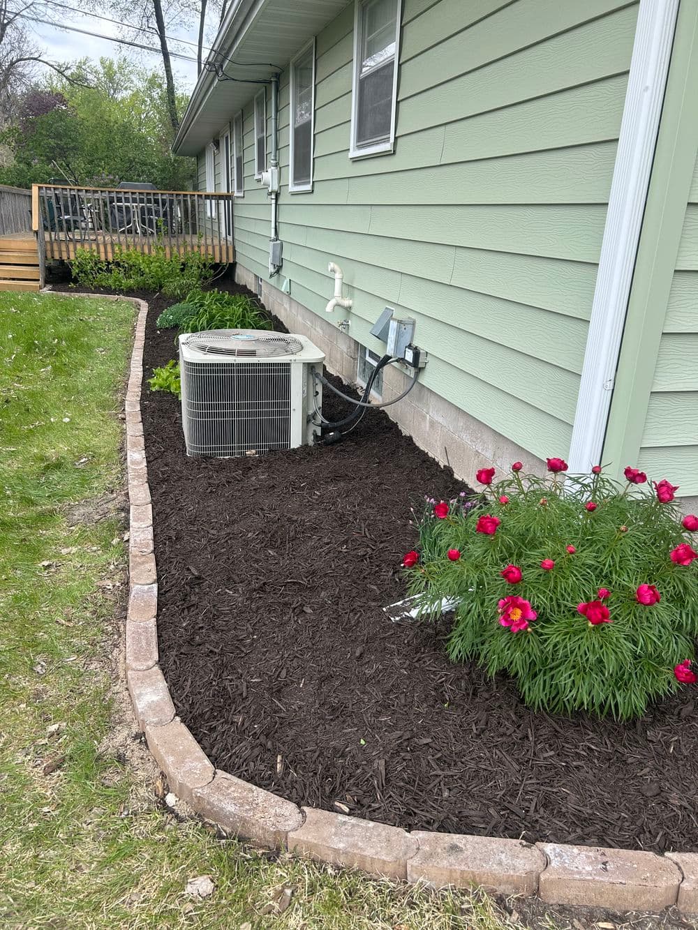 Air conditioning unit and flower bed with red peonies next to green house exterior.